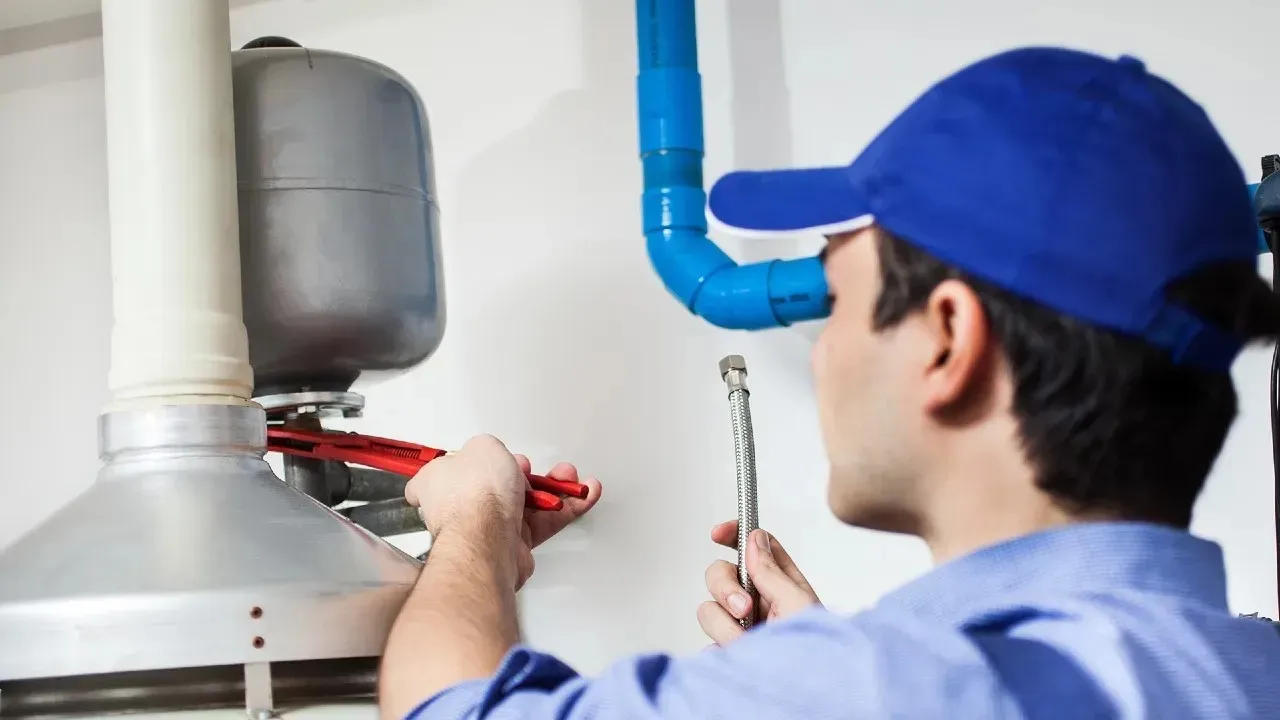 A plumber using a red pipe wrench to adjust an expansion tank on a water heater system while holding a braided hose.