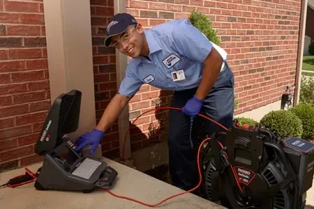A professional technician in a light blue uniform and purple gloves operating a digital monitor for an in-line video camera sewer inspection outside a brick house.