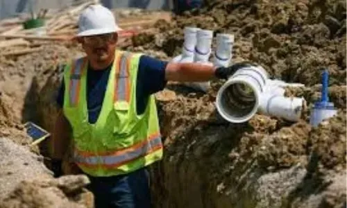 A construction plumber wearing a hard hat and safety vest standing in a trench next to large PVC sewer pipes.