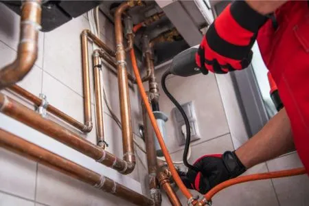 A professional technician in red and black safety gloves using a specialized electronic leak detection probe on a network of copper pipes.